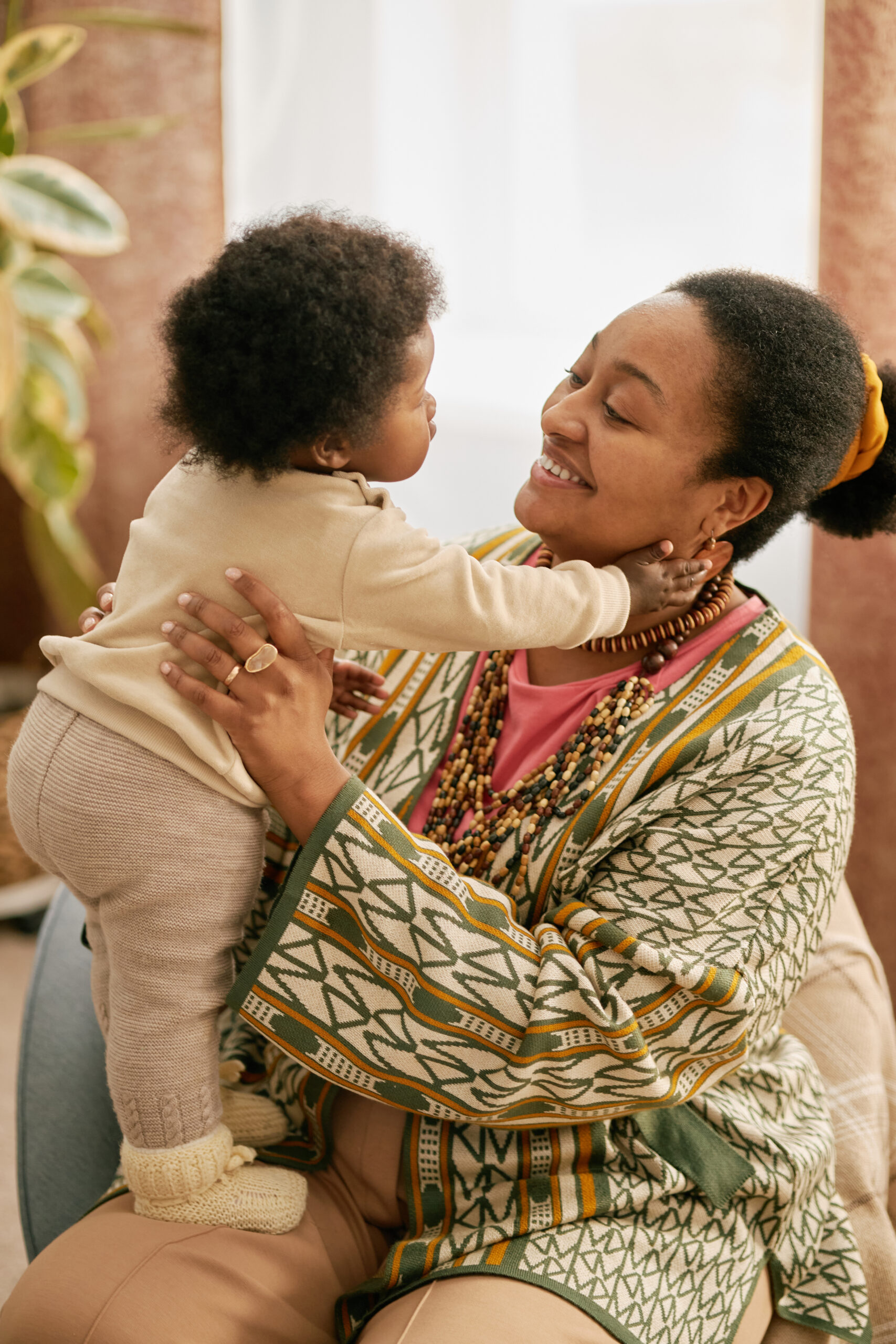 Vertical shot of smiling Black mother in patterned clothes playing with little baby holding child on laps in cozy home setting