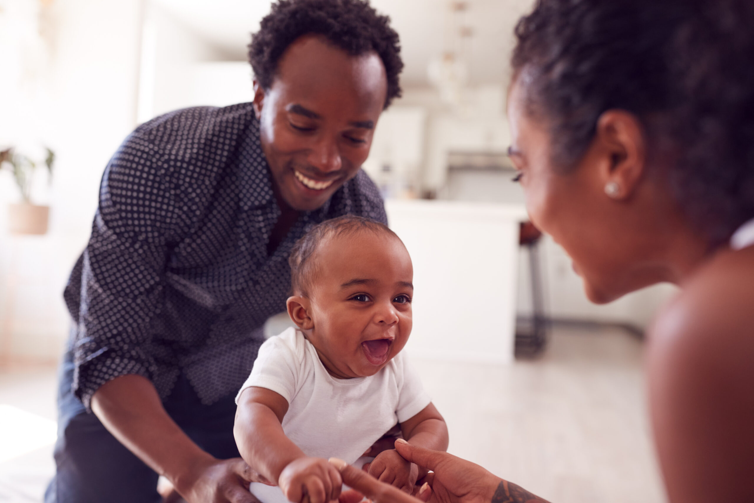 Parents Encouraging Smiling Baby Daughter To Take First Steps And Walk At Home