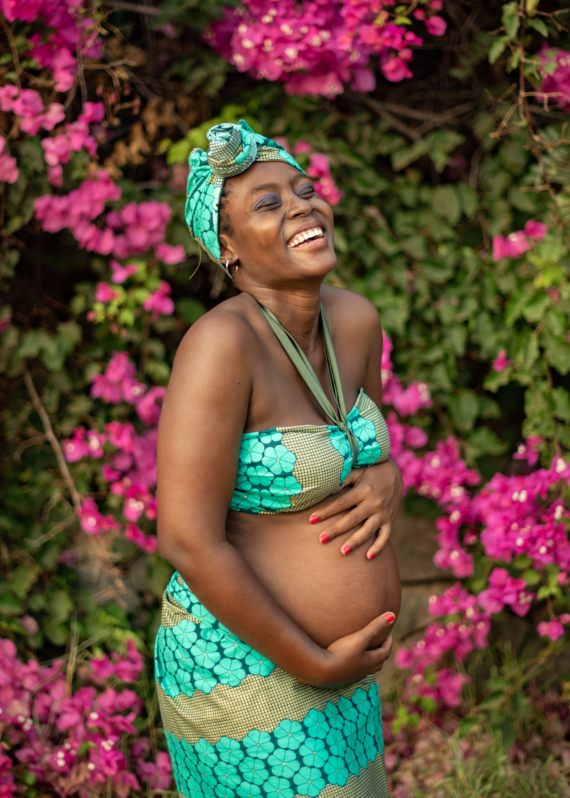 Cheerful pregnant black female in traditional clothes touching tummy and laughing while standing near blooming shrub in garden