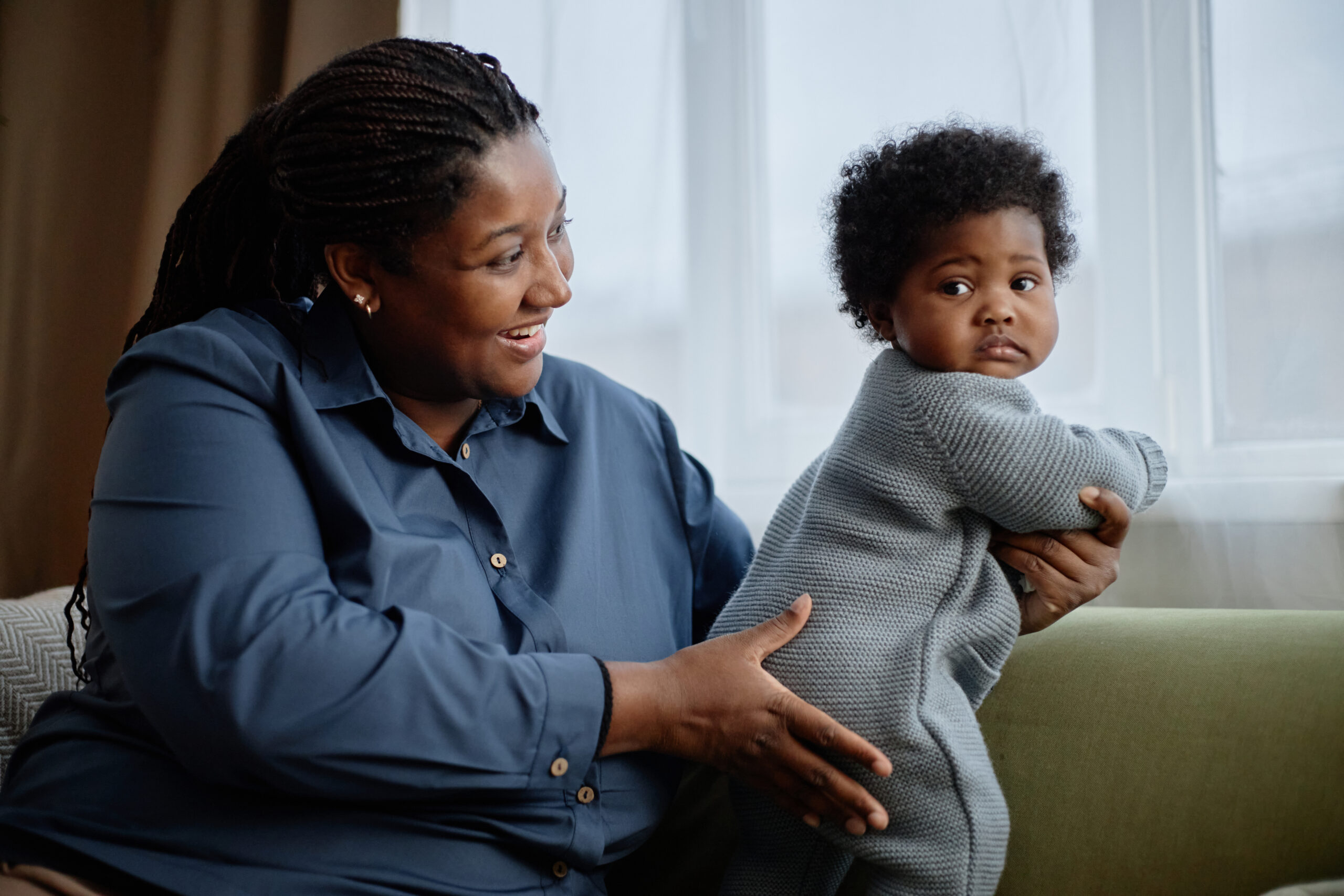 Medium shot of happy Black woman gently holding little baby girl dressed in comfy knitted romper of blue color standing up on sofa, captured against window at home