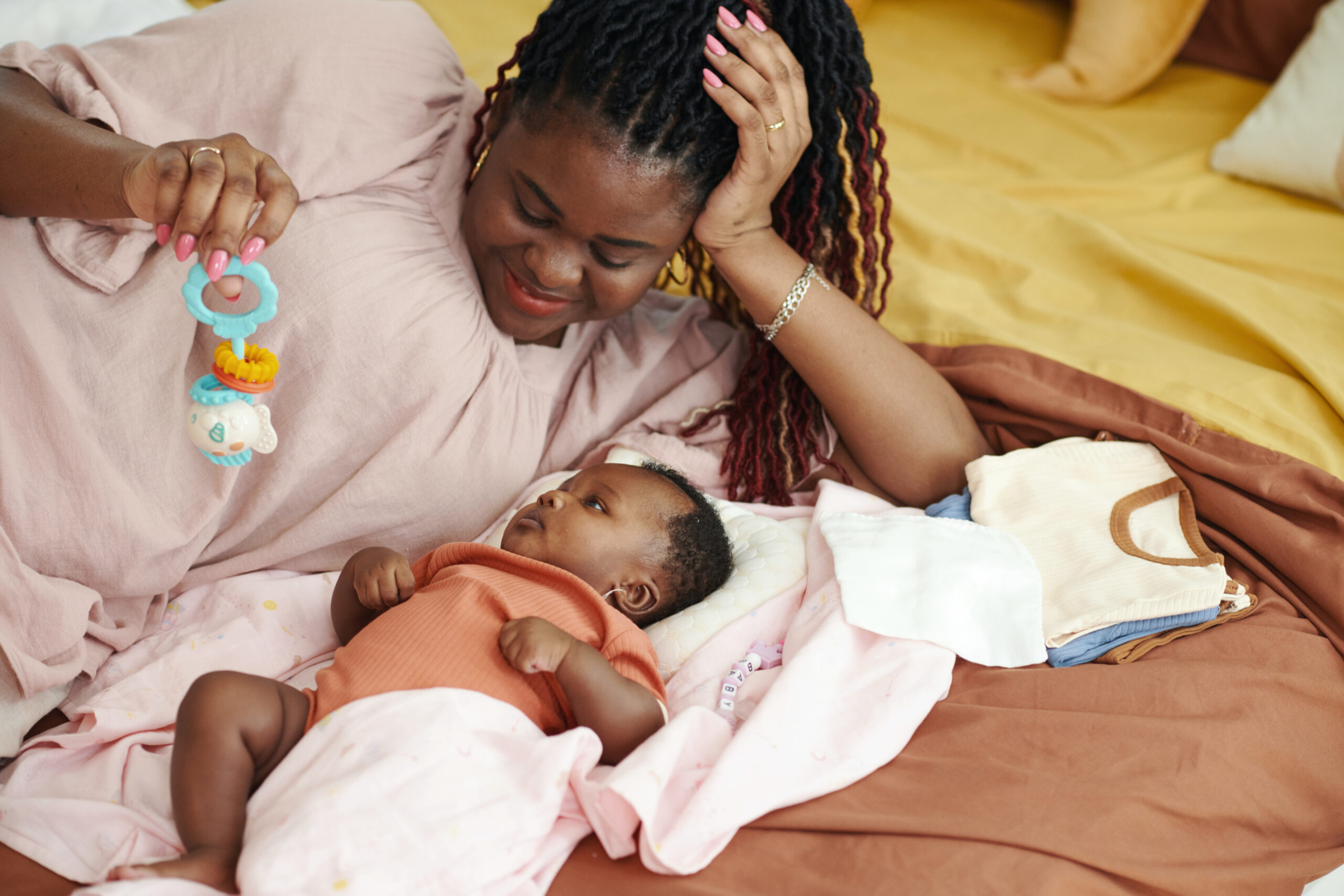 Happy mother lying in bed with newborn baby talking with her and showing rattles