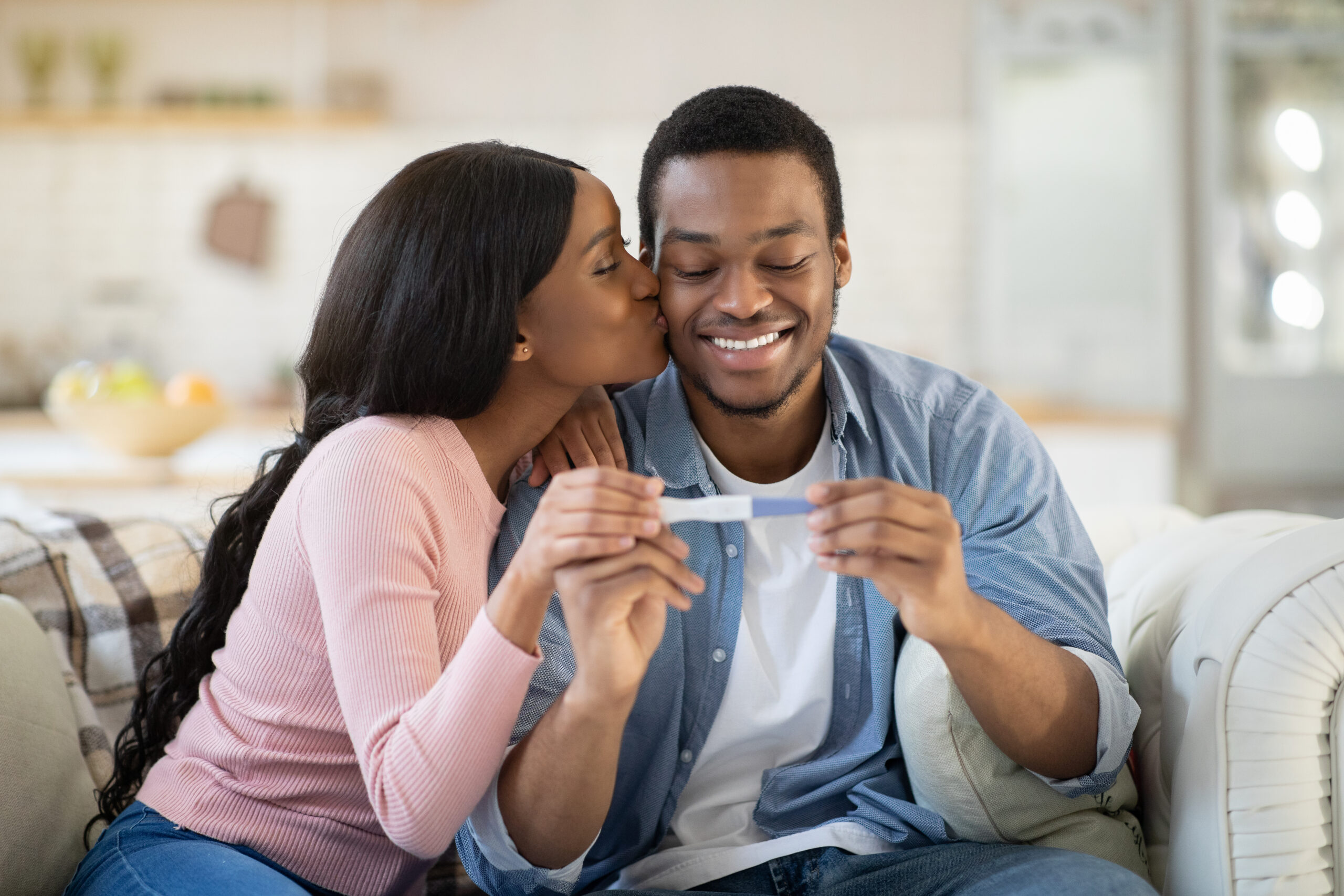 Attractive black woman kissing her husband with positive pregnancy test at home. African American couple happy to find out about their future baby, exchanging caresses indoors