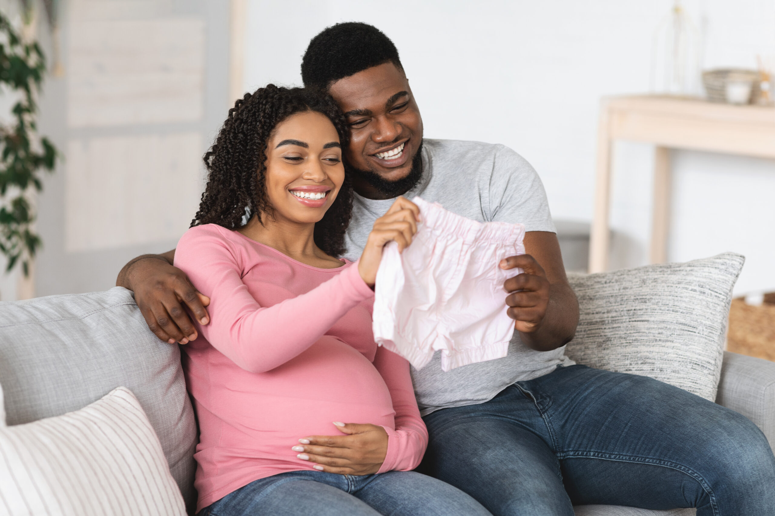Smiling black pregnant couple holding pink baby clothes, enjoying future parenthood, home interior, free space. Joyful african american parents looking at baby shorts, waiting for baby