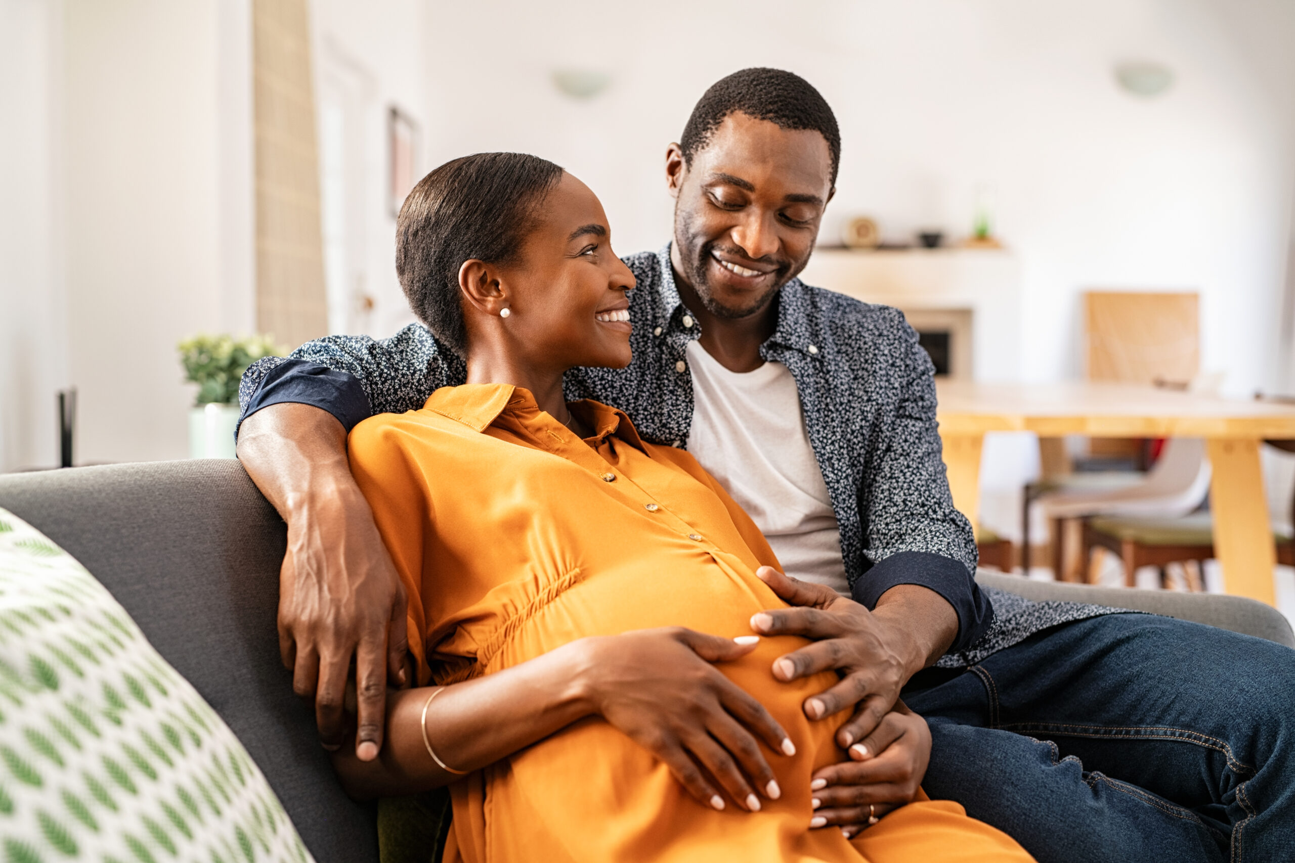 Happy mature loving husband and black pregnant woman embracing belly at home. Smiling middle aged african american man hugging pregnant woman sitting on couch holding tummy. Beautiful mid adult couple expecting baby and dreaming on sofa.