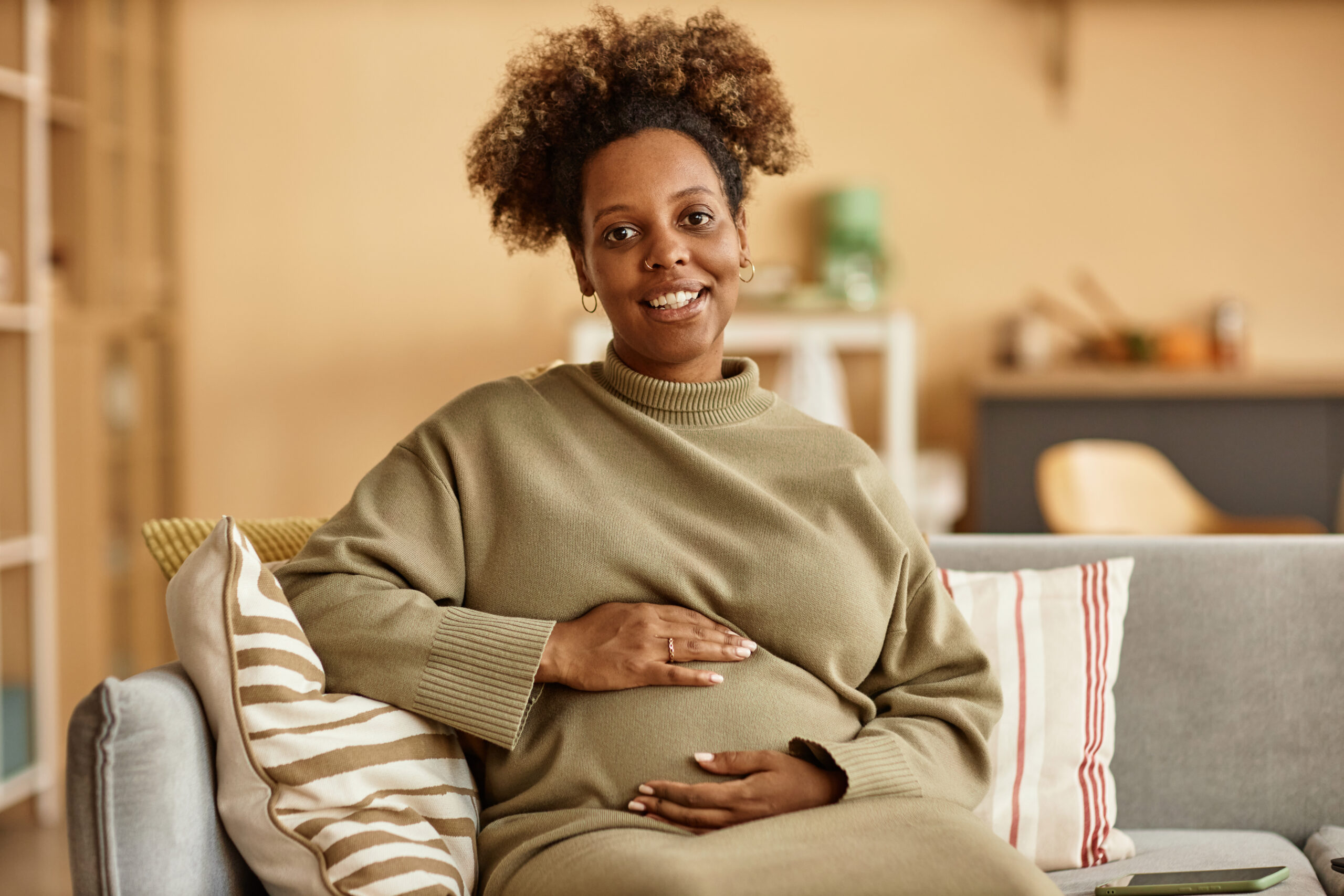 Smiling pregnant African American woman in pastel green sweater dress looking at camera while sitting on sofa at home embracing her round belly