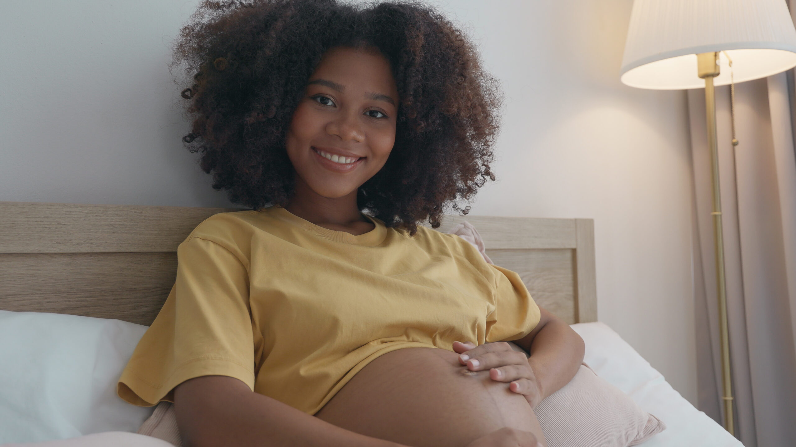 A beaming young woman with a full head of curly hair sits comfortably, cradling baby bump with tender care. The soft indoor lighting enhances the serene atmosphere as enjoys