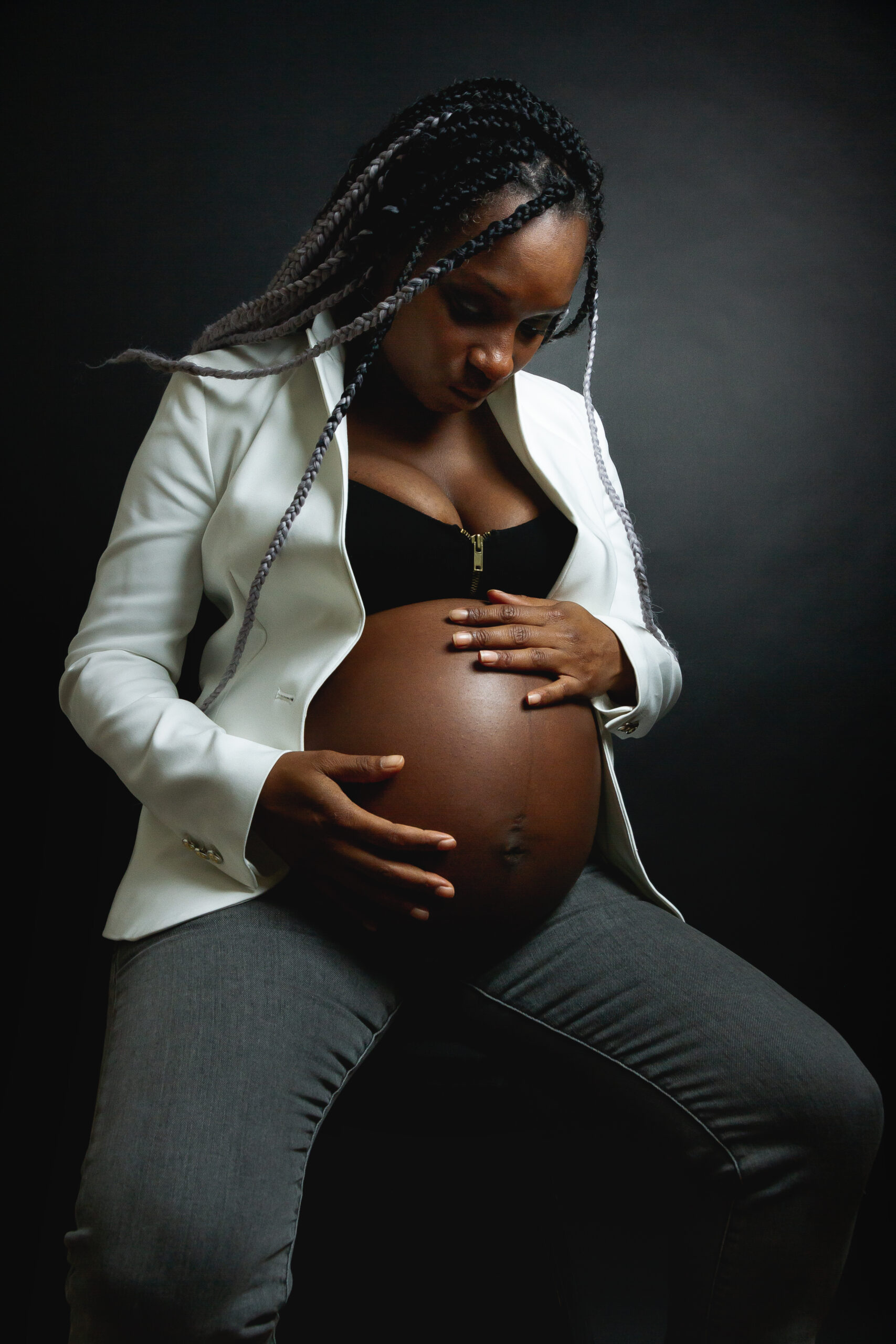 Young pregnant African American female with braids wearing white blazer and jeans stroking belly while sitting in studio against black wall