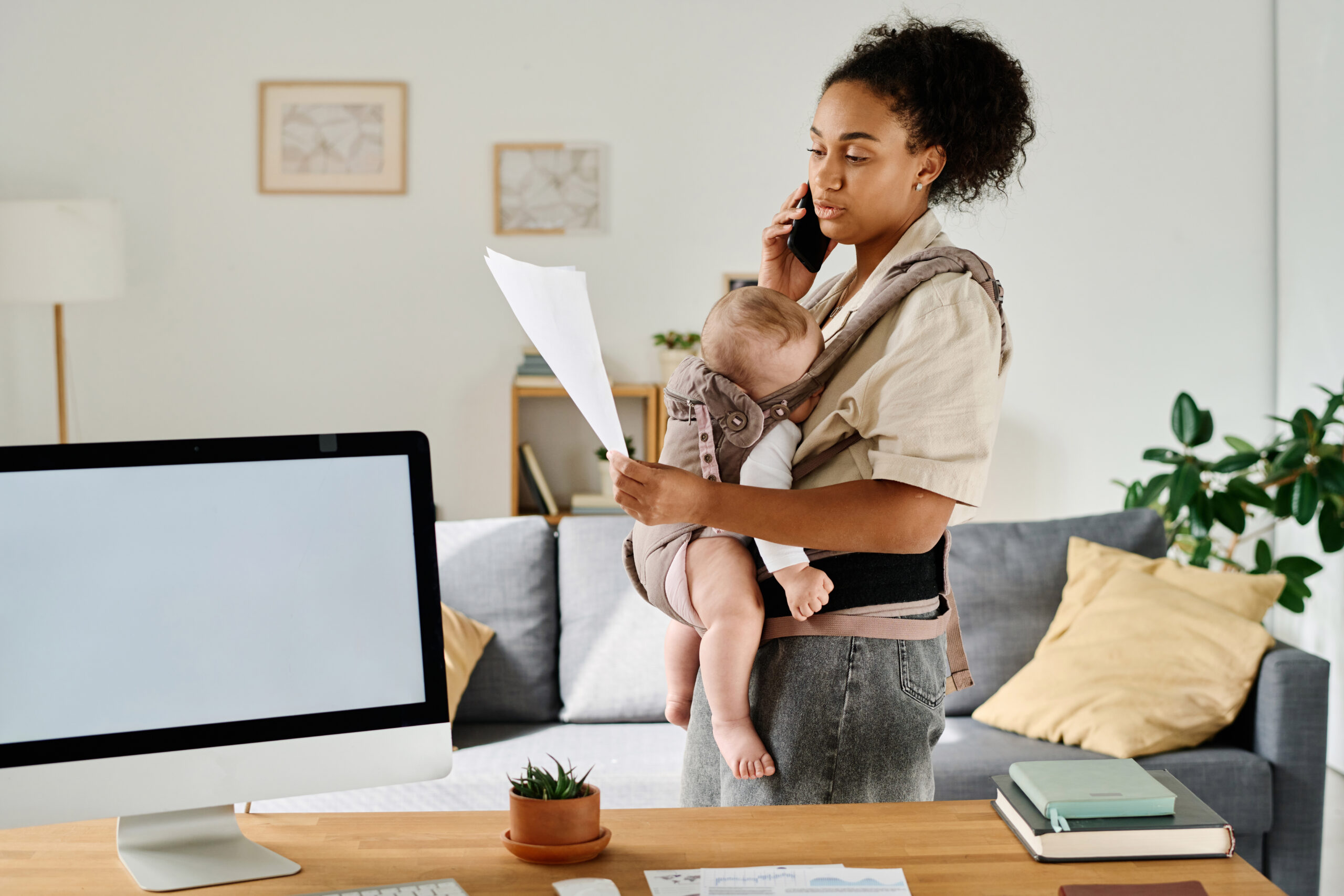 Young mom talking on the phone and reading document, she working at home while sitting with baby